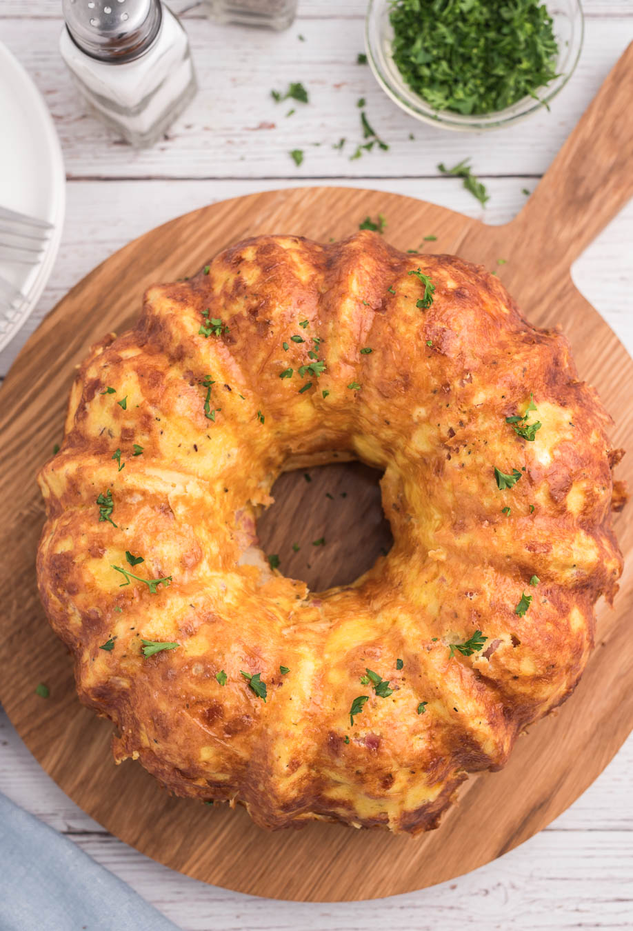 up close view of breakfast casserole shaped by bundt pan and placed on serving board with parsley for garnish. chopped parsley in small dish, salt and pepper shakers on side.