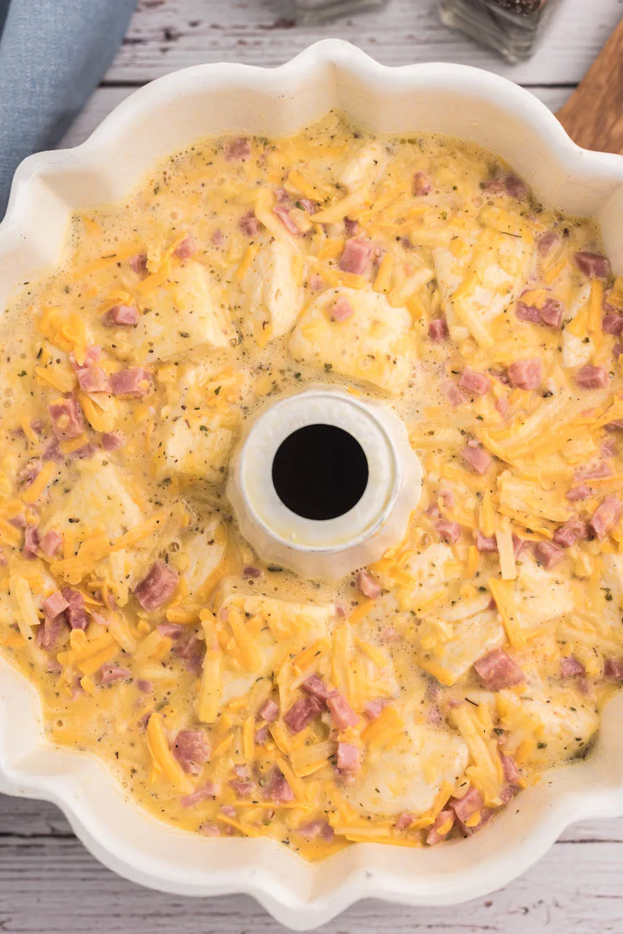 egg casserole mixture added to a bundt pan to prepare for cooking. ham bits, shredded cheese and biscuit chunks