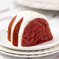 slice of a red bundt cake topped with cream cheese frosting on a stack of white plates