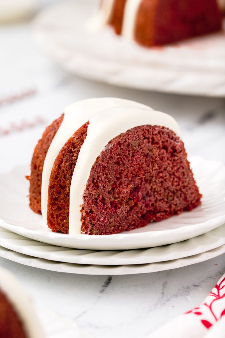 slice of a red bundt cake topped with cream cheese frosting on a stack of white plates