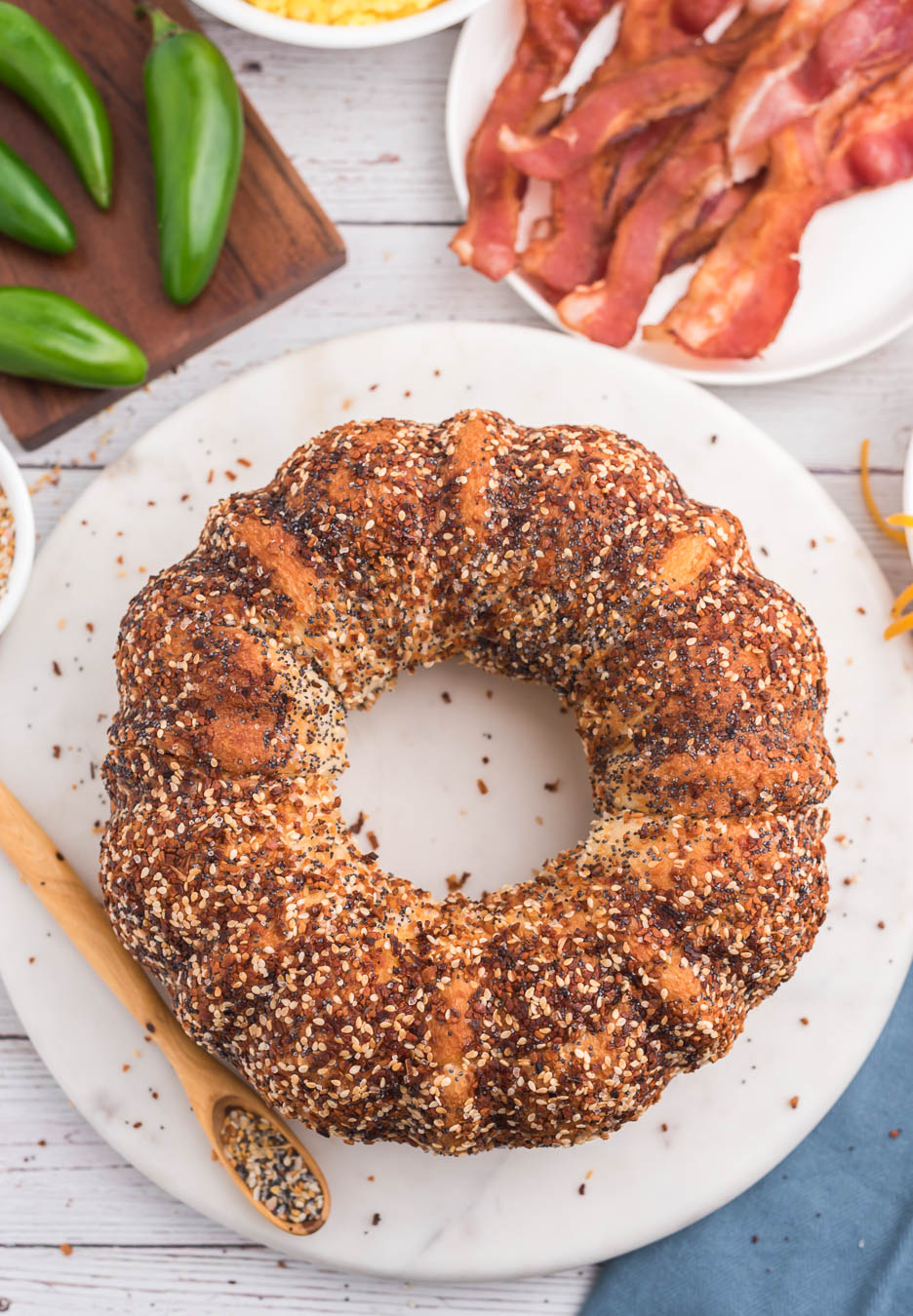 birds eye image of finished jumbo everything bagel on a marble cutting board with a wooden spoon next to it